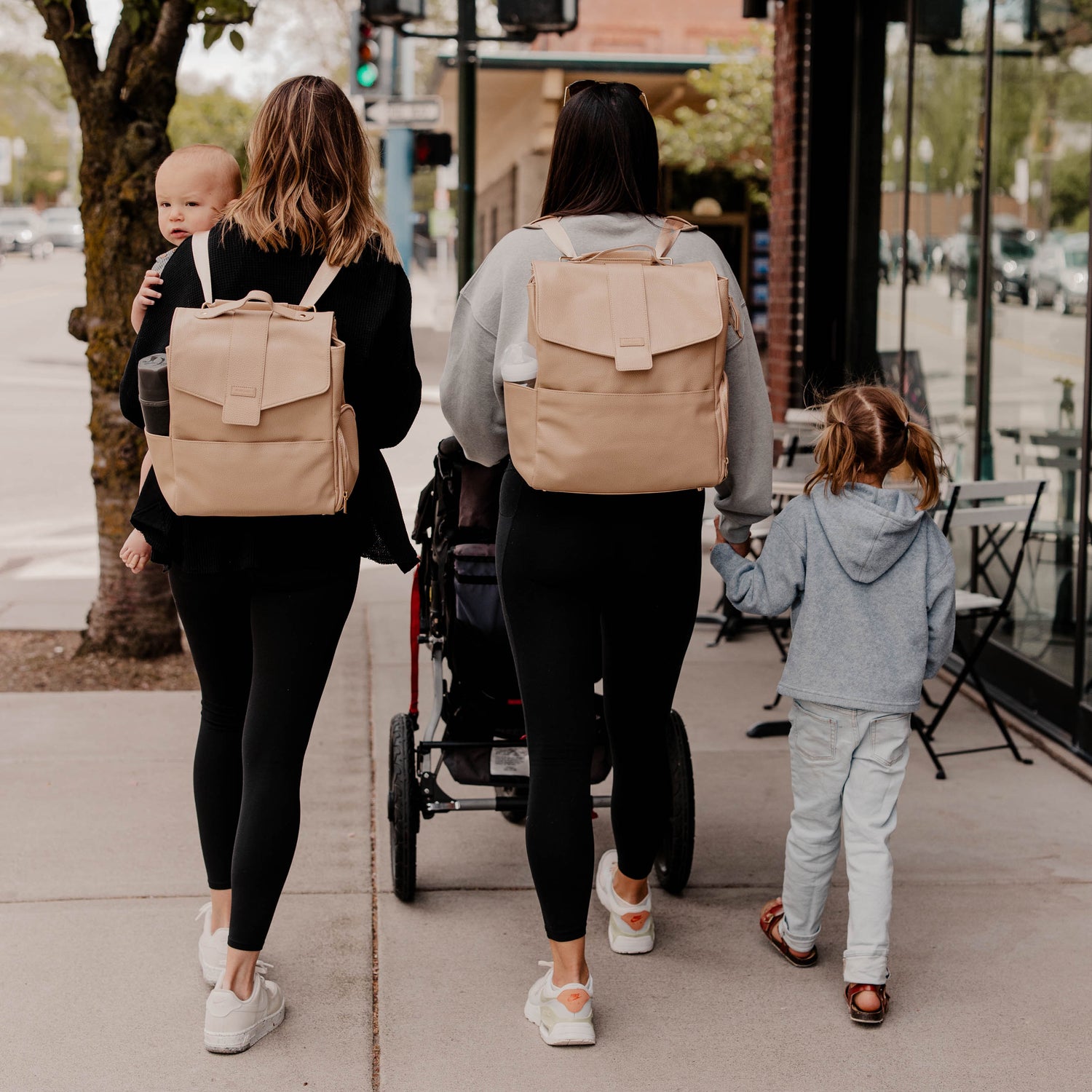Two women with beige backpacks walking with a baby and child on a sidewalk.